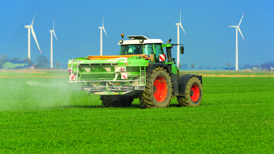 Tractor with fertilizer spreader on a field, windmills in the background. 