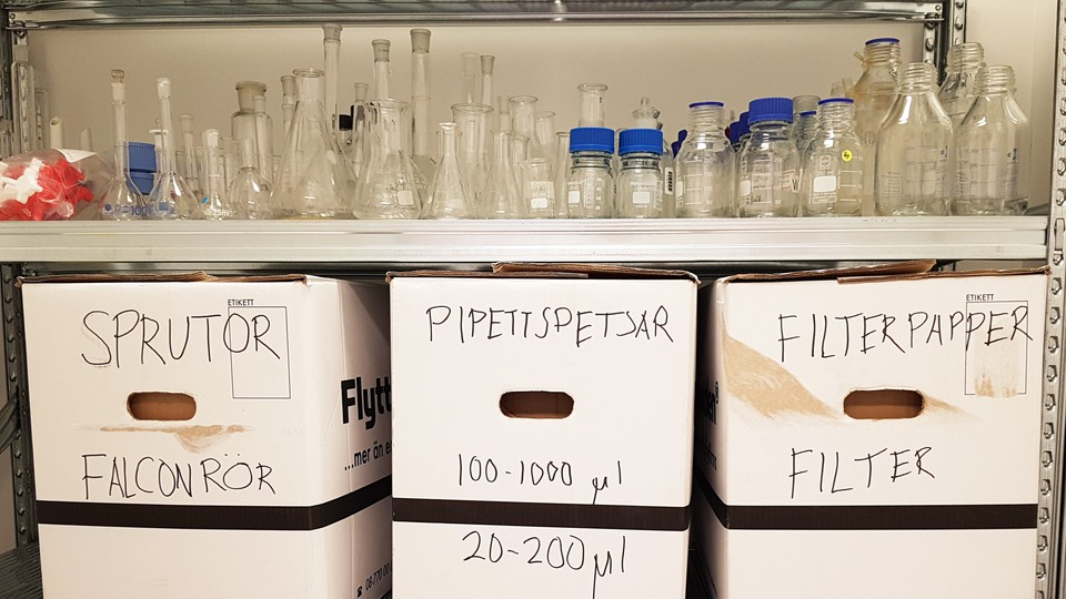 Lådor med orden sprutrör, pipettspetsar och filterpapper. På hyllan ovanför står olika glasflaskor och kolvar för använning i laboratorium.